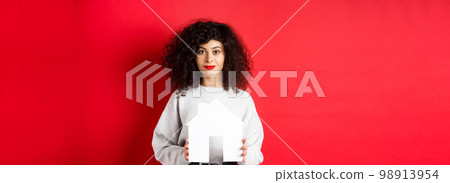 Real estate. Young caucasian woman in casual clothes showing paper house cutout, buying property or renting apartment, standing on red background Real estate. Young caucasian woman in casual clothes showing paper house cutout, buying property or renting apartment, standing on red background 98913954