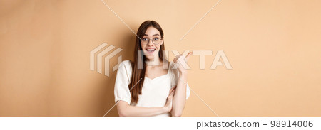 Excited girl in glasses pointing aside at logo, smiling amazed, recommending company, standing on beige background 98914006