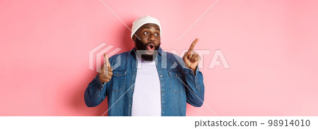 Happy african-american man showing thumb-up and pointing at upper left corner copy space, praising good deal, standing over pink background 98914010