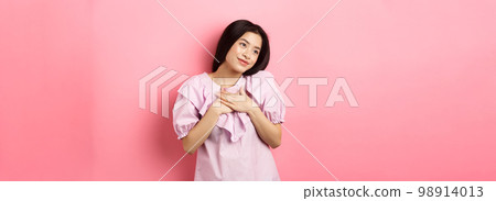 Dreamy asian girl smiling and holding hands on chest, looking left at something romantic and cute, standing on pink background Dreamy asian girl smiling and holding hands on chest, looking left at something romantic and cute, standing on pink background 98914013