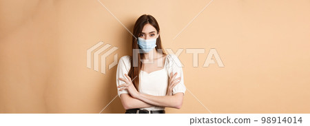 Covid-19 and lifestyle concept. Arrogant woman in medical mask frowning, cross arms on chest and looking confident at camera, beige background Covid-19 and lifestyle concept. Arrogant woman in medical mask frowning, cross arms on chest and looking confident at camera, beige background 98914014