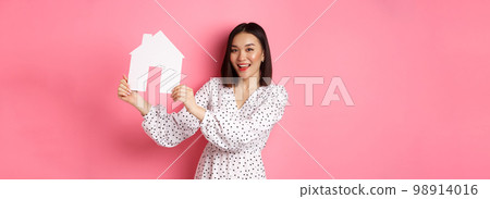 Real estate. Beautiful asian woman demonstrating paper house model, looking at camera confident, advertising home for sale, standing over pink background 98914016