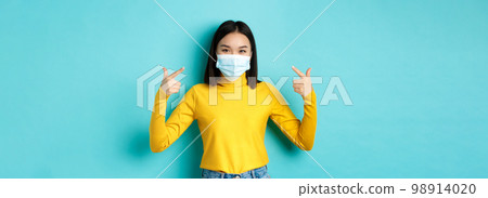 Covid-19, social distancing and pandemic concept. Young asian woman protect herself from coronavirus, pointing finger at her medical mask, standing over blue background Covid-19, social distancing and pandemic concept. Young asian woman protect herself from coronavirus, pointing finger at her medical mask, standing over blue background 98914020