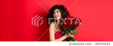 Tender young woman in elegant red dress, holding romantic bouquet of red roses and looking right, smiling dreamy, thinking about lover on Valentines day, studio background Tender young woman in elegant red dress, holding romantic bouquet of red roses and looking right, smiling dreamy, thinking about lover on Valentines day, studio background 98914045