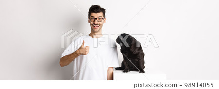 Cheerful handsome guy standing near cute black pug and showing thumb-up. Pet owner approve and recommend product for dogs, white background 98914055
