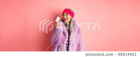 Shopping and fashion concept. Stylish asian senior woman in trendy clothes showing plastic credit card, smiling sassy, standing over pink background 98914081
