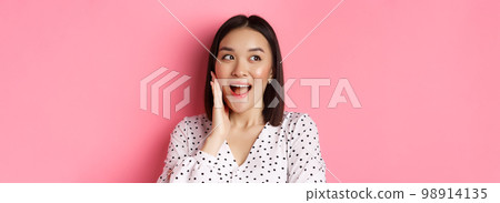 Coquettish asian woman in blushing, touching cheek and looking left amused, standing over pink background 98914135