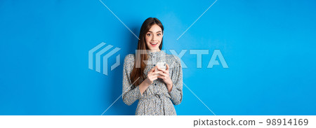 Cute smiling girl with long natural hair, wearing dress, using smartphone and looking happy, standing against blue background Cute smiling girl with long natural hair, wearing dress, using smartphone and looking happy, standing against blue background 98914169
