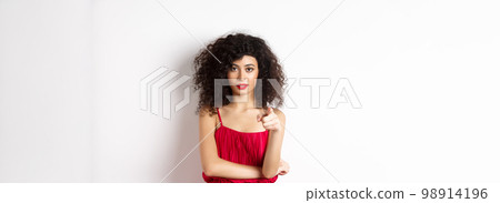 Confident young woman in red dress and makeup, pointing at camera, choosing you, inviting to event, standing over white background 98914196