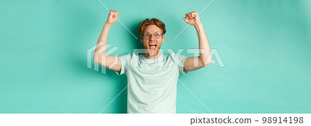 Young redhead man feeling like champion, raising hands up in fist pump gesture and shouting yes with joy, winning prize, triumphing of success, standing over mint background 98914198