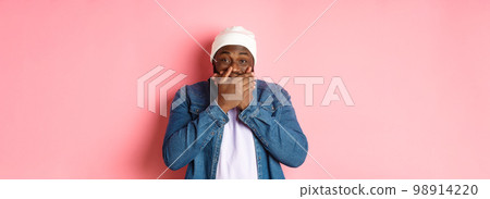 Happy Black hipster man holding laugh, cover mouth and giggle over funny joke, staring at camera and chuckling, standing over pink background Happy Black hipster man holding laugh, cover mouth and giggle over funny joke, staring at camera and chuckling, standing over pink background 98914220