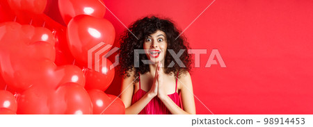 Valentines day. Excited caucasian woman in dress jumping from amazement, looking at something taunting, want to get product, standing near hearts balloons, white background 98914453
