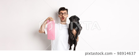 Image of hipster guy pet owner, holding cute black pug and dog poop bag, standing over white background 98914637