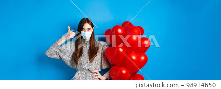 Coronavirus and pandemic concept. Beautiful woman in medical mask and dress standing near Valentines day balloons and showing thumb up, standing on blue background Coronavirus and pandemic concept. Beautiful woman in medical mask and dress standing near Valentines day balloons and showing thumb up, standing on blue background 98914650