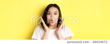Close up of surprised asian girl say wow, stare at camera amzed with hands near face, standing over yellow background 98914672