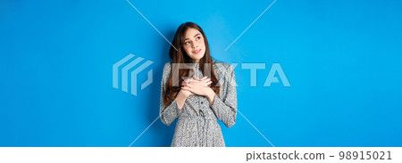 Romantic dreamy girl in dress looking at upper left corner, holding hands on heart and daydreaming, imaging something cute, standing on blue background Romantic dreamy girl in dress looking at upper left corner, holding hands on heart and daydreaming, imaging something cute, standing on blue background 98915021