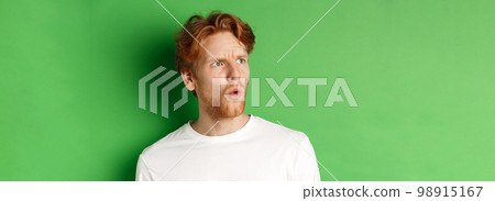 Confused redhead man staring left and looking worried, standing against green background 98915167