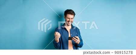 Happy young man winning in video game on mobile phone, looking satisfied at smartphone screen and say yes with joy, standing on blue background Happy young man winning in video game on mobile phone, looking satisfied at smartphone screen and say yes with joy, standing on blue background 98915278