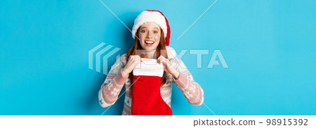 Beautiful redhead girl in santa hat open Christmas stocking and looking surprised, receiving xmas gift, standing over blue background Beautiful redhead girl in santa hat open Christmas stocking and looking surprised, receiving xmas gift, standing over blue background 98915392