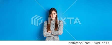 Angry frowning woman in dress, cross arms on chest and feel offended about something unfair, standing upset against blue background Angry frowning woman in dress, cross arms on chest and feel offended about something unfair, standing upset against blue background 98915413