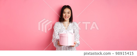 Beautiful asian woman wishing happy holidays, giving you gift in cute box, standing against pink background 98915415