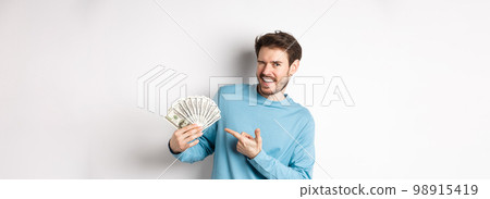 Handsome rich guy winking at camera, showing income, pointing finger at earned money and smiling, standing over white background 98915419