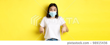 Covid-19, pandemic and social distancing concept. Amazed asian woman in medical mask, showing advertisement, pointing right and smiling, yellow background 98915436