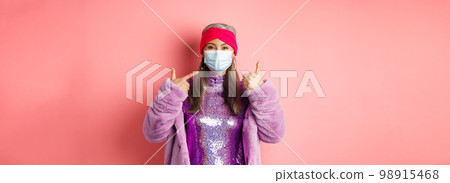 Covid-19, social distancing and fashion concept. Smiling asian senior woman in fashionable clothes and respirator, pointing at face mask and show thumb-up, pink background Covid-19, social distancing and fashion concept. Smiling asian senior woman in fashionable clothes and respirator, pointing at face mask and show thumb-up, pink background 98915468