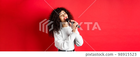 Leisure and hobbies concept. Stylish young woman singing karaoke, looking aside and holding microphone, performing song, standing on red background Leisure and hobbies concept. Stylish young woman singing karaoke, looking aside and holding microphone, performing song, standing on red background 98915523