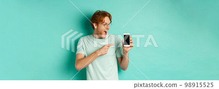 Check this out. Handsome redhead guy in glasses pointing finger at blank smartphone screen, showing online promotion, standing amazed over turquoise background Check this out. Handsome redhead guy in glasses pointing finger at blank smartphone screen, showing online promotion, standing amazed over turquoise background 98915525
