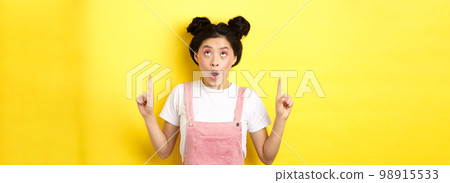 Excited asian teen student with glamour makeup, gasping amazed pointing fingers up, showing advertisement, yellow background 98915533