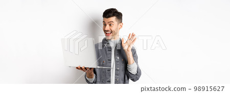 Excited man waiving hand at laptop, video chatting on computer and smiling friendly, standing on white background Excited man waiving hand at laptop, video chatting on computer and smiling friendly, standing on white background 98915627