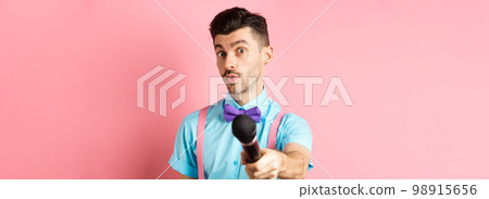 Handsome male show host stretch out hand with microphone, asking for interview or comments, standing on pink background 98915656