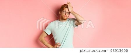 Tired young man with red hair, wearing glasses, looking annoyed and tensed, making facepalm gesture and exhale bothered, standing over pink background 98915696