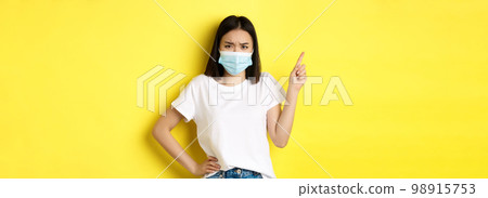 Covid, health care and pandemic concept. Asian female model in medical mask and white t-shirt pointing finger at upper left corner logo, showing promotion, yellow background 98915753