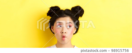 Close-up of teen asian girl pucker lips and looking funny at camera, standing with glamour makeup and stylish hairstyle, yellow background 98915756