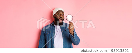 Happy african-american man looking through magnifying glass, smiling amazed, standing against pink background 98915801