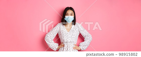 Covid-19, quarantine and lifestyle concept. Cute korean woman in dress and face mask using preventive measures from coronavirus, standing over pink background Covid-19, quarantine and lifestyle concept. Cute korean woman in dress and face mask using preventive measures from coronavirus, standing over pink background 98915802