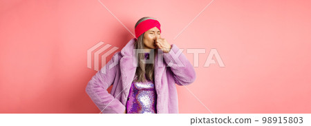 Portrait of stylish old asian woman in fashionable purple coat and dress, shut nose from bad smell, express disgust and aversion as something sink awfully, standing over pink background Portrait of stylish old asian woman in fashionable purple coat and dress, shut nose from bad smell, express disgust and aversion as something sink awfully, standing over pink background 98915803