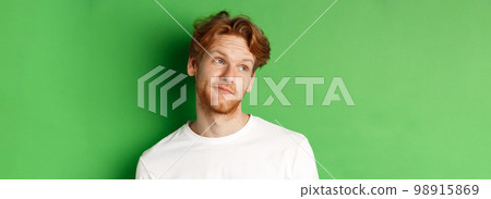 Smiling redhead man with messy haircut and beard tilt head, looking left with pleased face, standing over green background 98915869