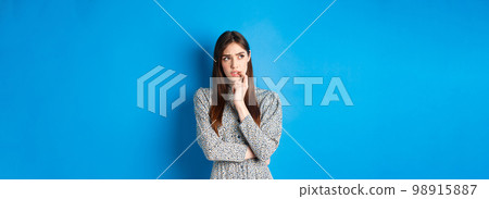 Troubled pensive girl looking aside at logo, biting fingernail and thinking, standing on blue background in long dress 98915887