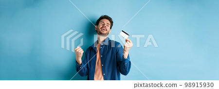 Happy and satisfied young man dancing with plastic credit card, fist pump and smile, standing on blue background Happy and satisfied young man dancing with plastic credit card, fist pump and smile, standing on blue background 98915930