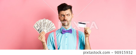 Doubtful and indecisive guy looking hesitant at plastic credit card, holding cash dollars, standing over pink background Doubtful and indecisive guy looking hesitant at plastic credit card, holding cash dollars, standing over pink background 98915932