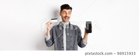 Handsome smiling man in denim jacket looking at empty smartphone screen and showing plastic credit card, good internet offer, standing against white background Handsome smiling man in denim jacket looking at empty smartphone screen and showing plastic credit card, good internet offer, standing against white background 98915933