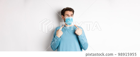 Coronavirus, health and quarantine concept. Confused young man pointing at medical mask on face and looking left, standing over white background 98915955