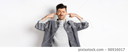 Annoyed young man plug ears with fingers and grimacing from loud noise, disturbed by noisy neighbours, standing at loud place on white background 98916017