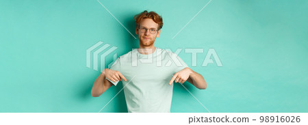 Skeptical and bothered man with ginger hair and beard, wearing glasses and t-shirt, smirk and point fingers down, showing promo with judgy face, turquoise background 98916026