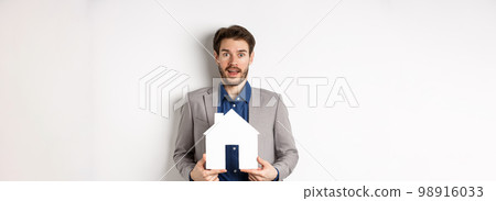 Real estate. Excited business man in suit holding paper house cutout and looking amazed at camera, buying apartment, white background Real estate. Excited business man in suit holding paper house cutout and looking amazed at camera, buying apartment, white background 98916033