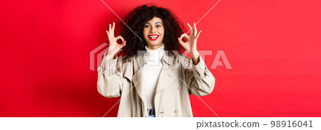 Stylish caucasian woman in trench coat showing okay gesture and smiling satisfied, recommending company, standing on red background 98916041