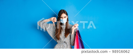 Covid-19, pandemic and lifestyle concept. Angry client in medical mask showing thumb down in dislike, frowning upset, holding shopping bags, blue background Covid-19, pandemic and lifestyle concept. Angry client in medical mask showing thumb down in dislike, frowning upset, holding shopping bags, blue background 98916062